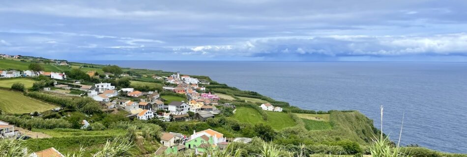 View over Ajuda da Bretanha on Sao Miguel, Azores. The foreground shows green grass with purple flowers at the background is the city with colorful buildings and the coastline and ocean behind it.