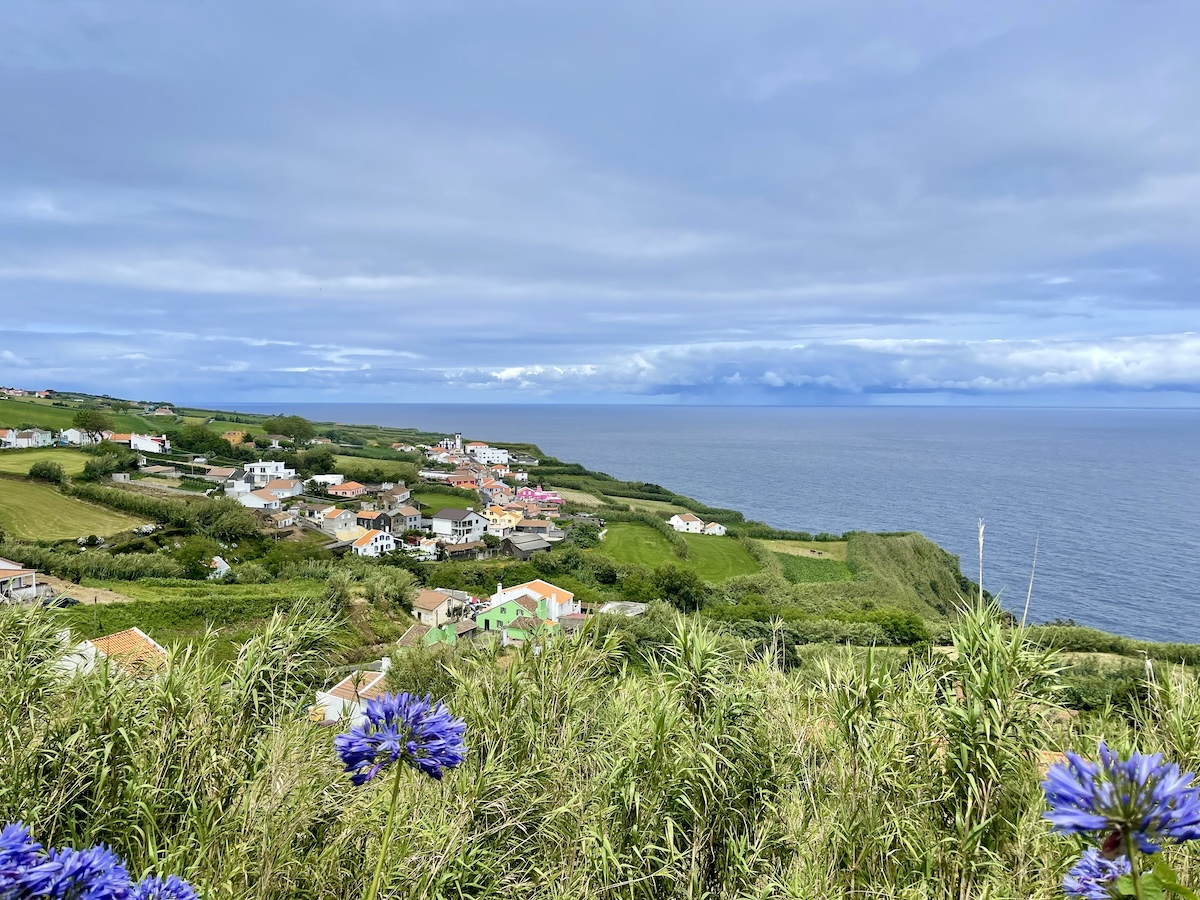 View over Ajuda da Bretanha on Sao Miguel, Azores. The foreground shows green grass with purple flowers at the background is the city with colorful buildings and the coastline and ocean behind it.