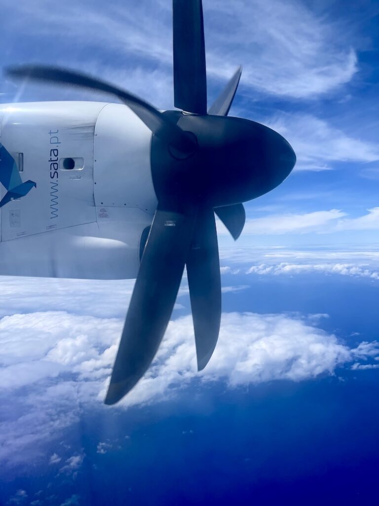 View out the window while flying over the Azores. The view is of the propeller of a SATA airplane with the backdrop of a blue sky with white clouds. 